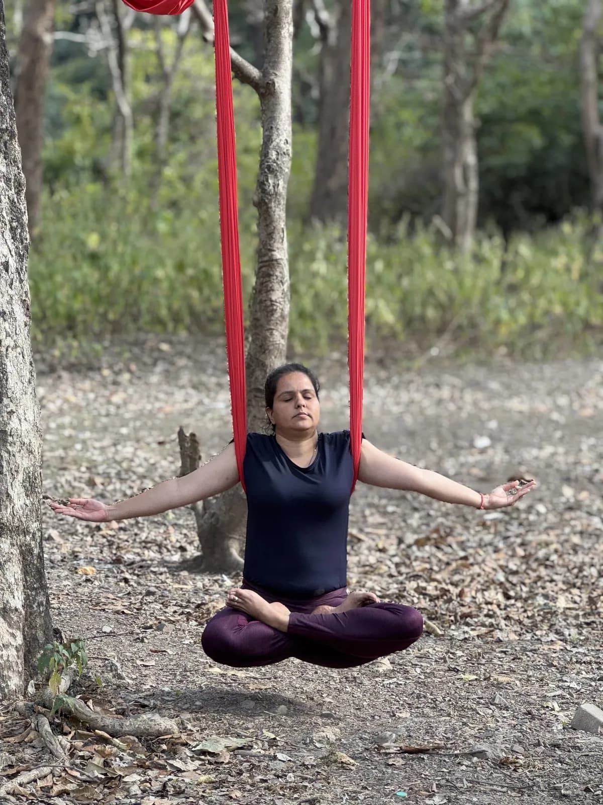 Student meditating cross-legged in lotus while floating in a red aerial hammock in a forest near Rishikesh