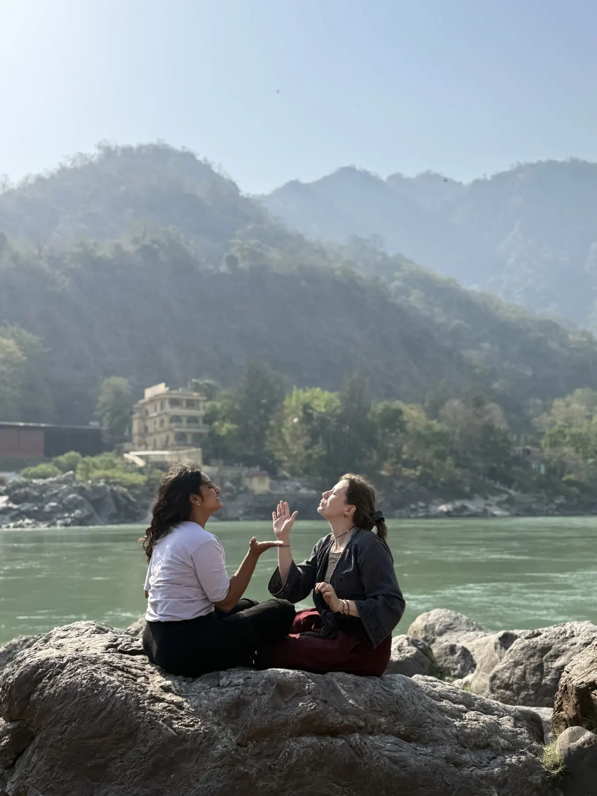 Two friends sitting on a rock by the turquoise Ganga with the forested Rishikesh hills behind