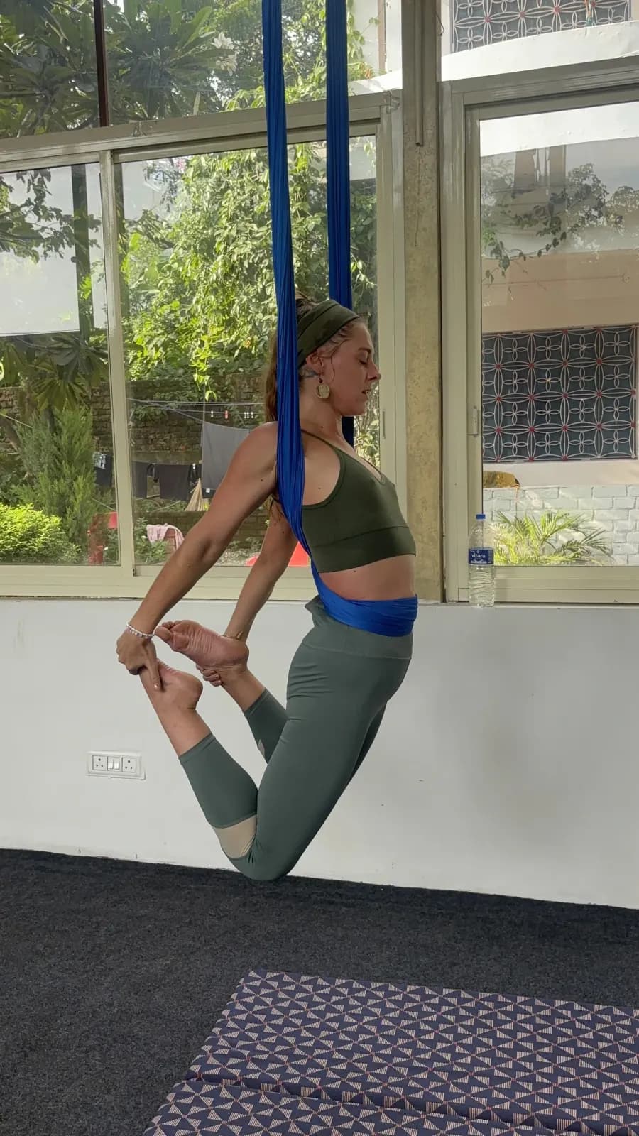 Student balancing in aerial dancer's pose with a blue aerial yoga hammock at her hips, holding her back foot, beside the studio window
