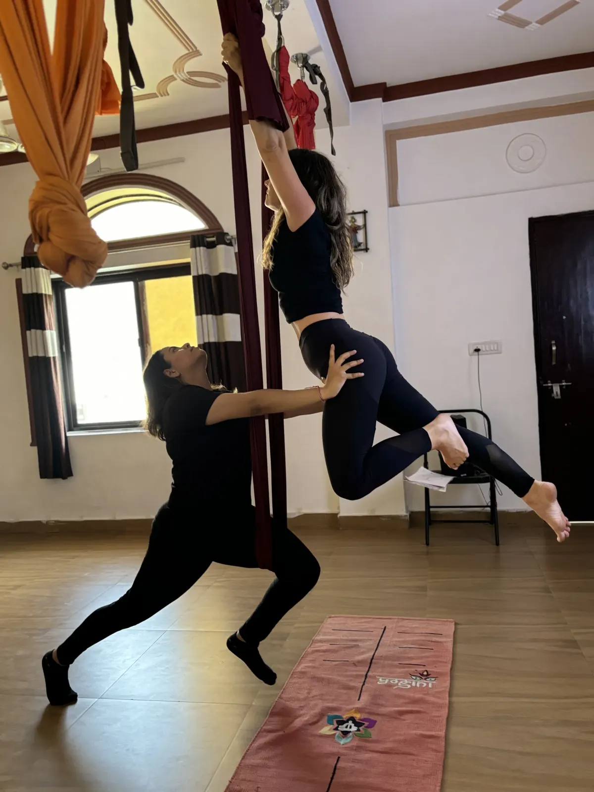 Aerial yoga teacher supporting a student's legs as she pulls herself up into the aerial silk in the Rishikesh studio