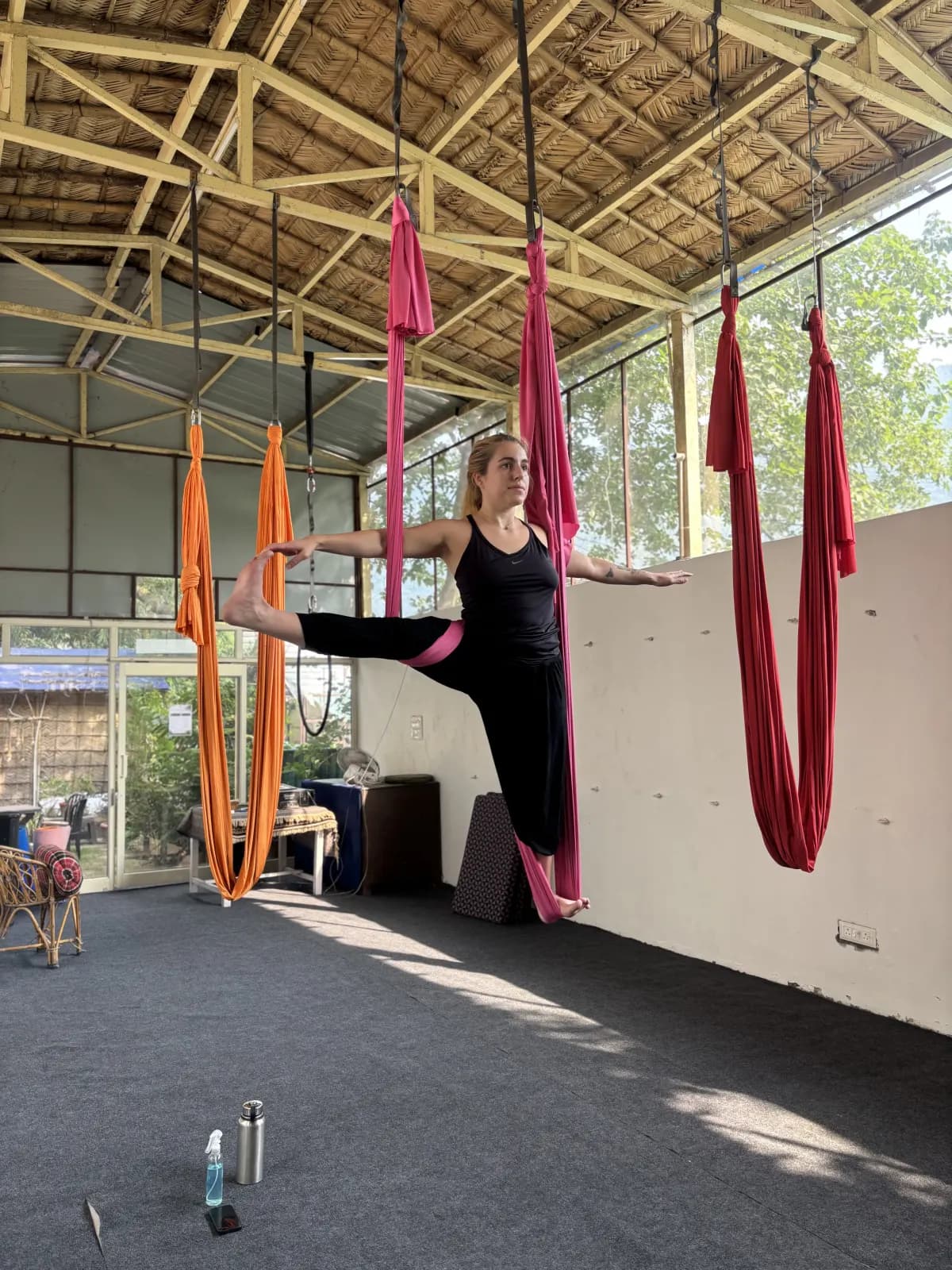 Student balancing in an aerial standing split with a pink hammock under her hip in the open-air bamboo-roof aerial yoga shala in Rishikesh