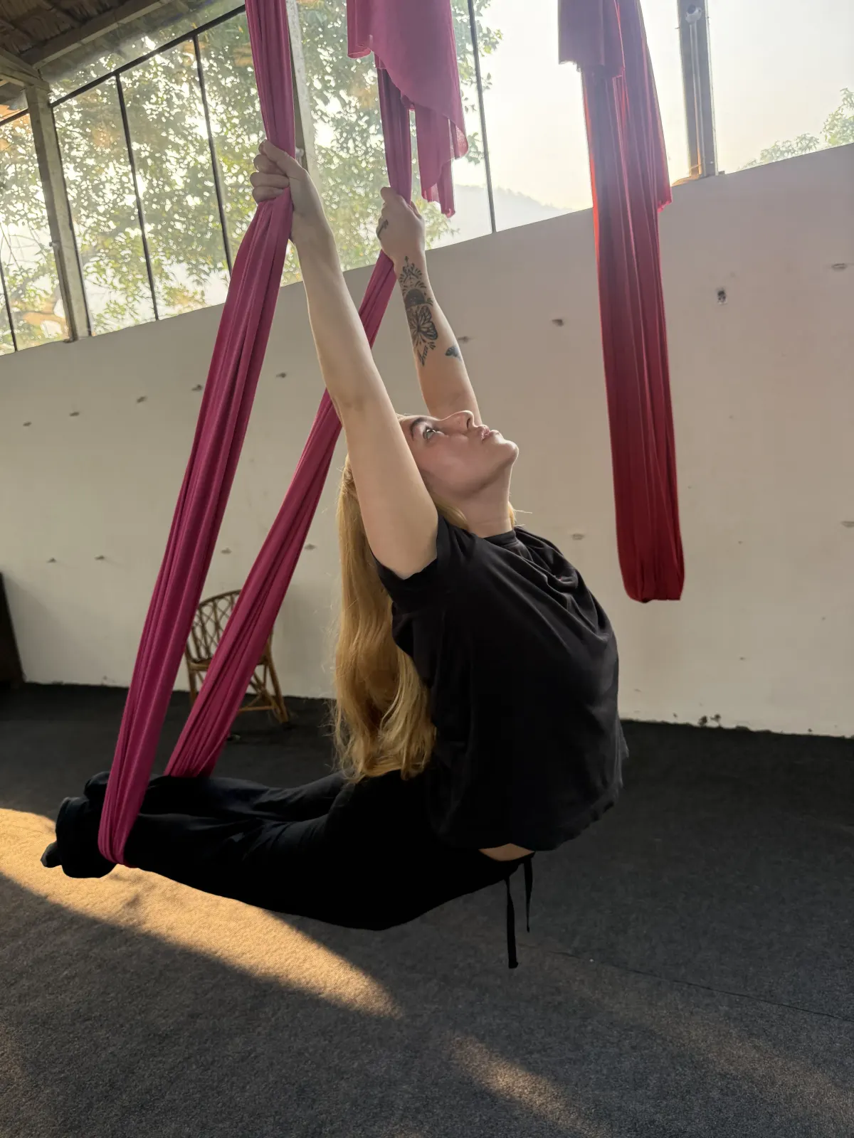 Student arching back into a supported aerial backbend with her legs wrapped in a maroon hammock, lit by morning sunlight