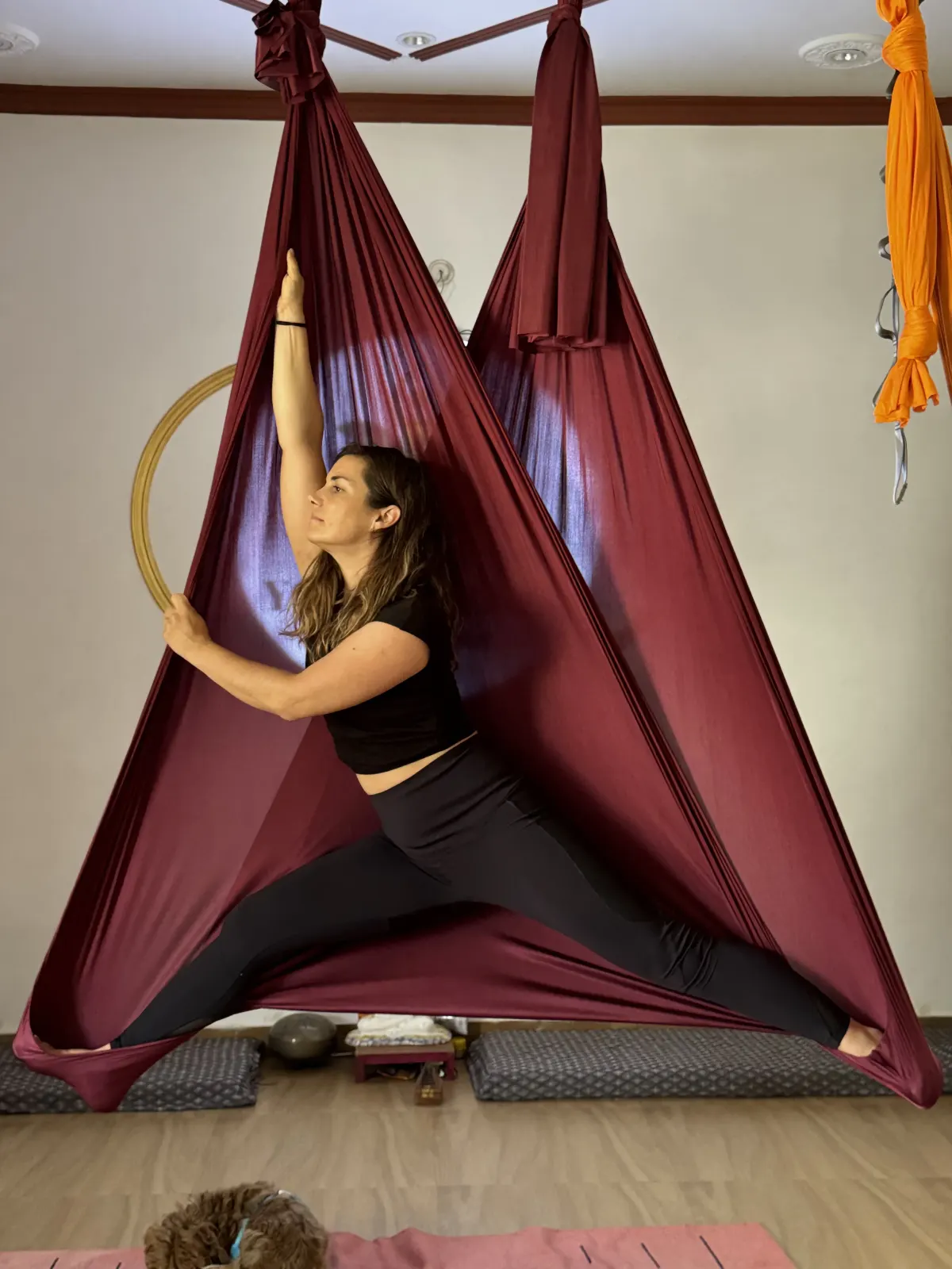 Student in a wide-leg aerial straddle with the maroon silk spread into a triangle in front of the Swaastik Yog School logo, Rishikesh