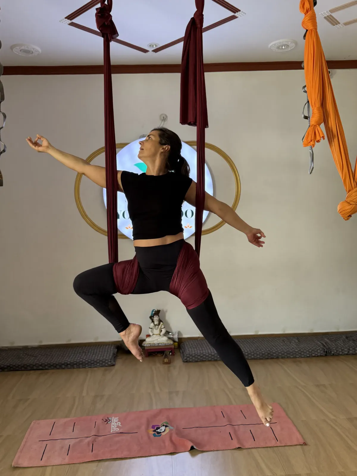 Student seated in the aerial hammock sling with one leg reaching to the floor and arms extended in a warrior-inspired aerial pose