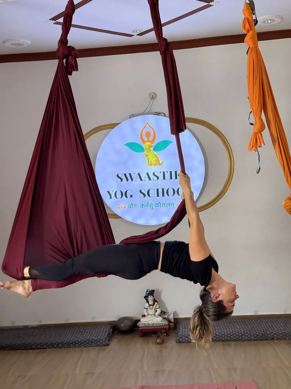 Student hanging upside down in an inverted aerial backbend, feet hooked in a maroon hammock, at Swaastik Yog School in Rishikesh
