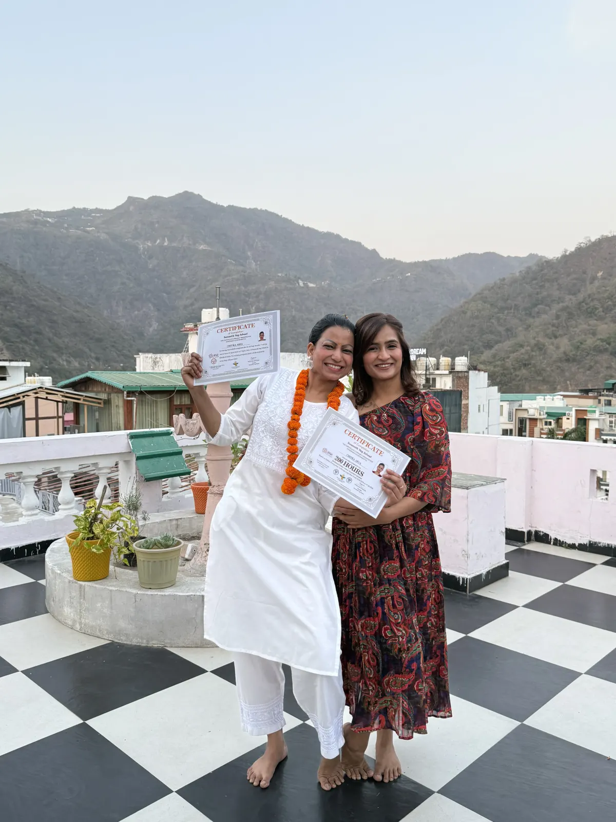 Two garlanded women holding Yoga Alliance teacher training certificates on the rooftop terrace with the Rishikesh hills behind