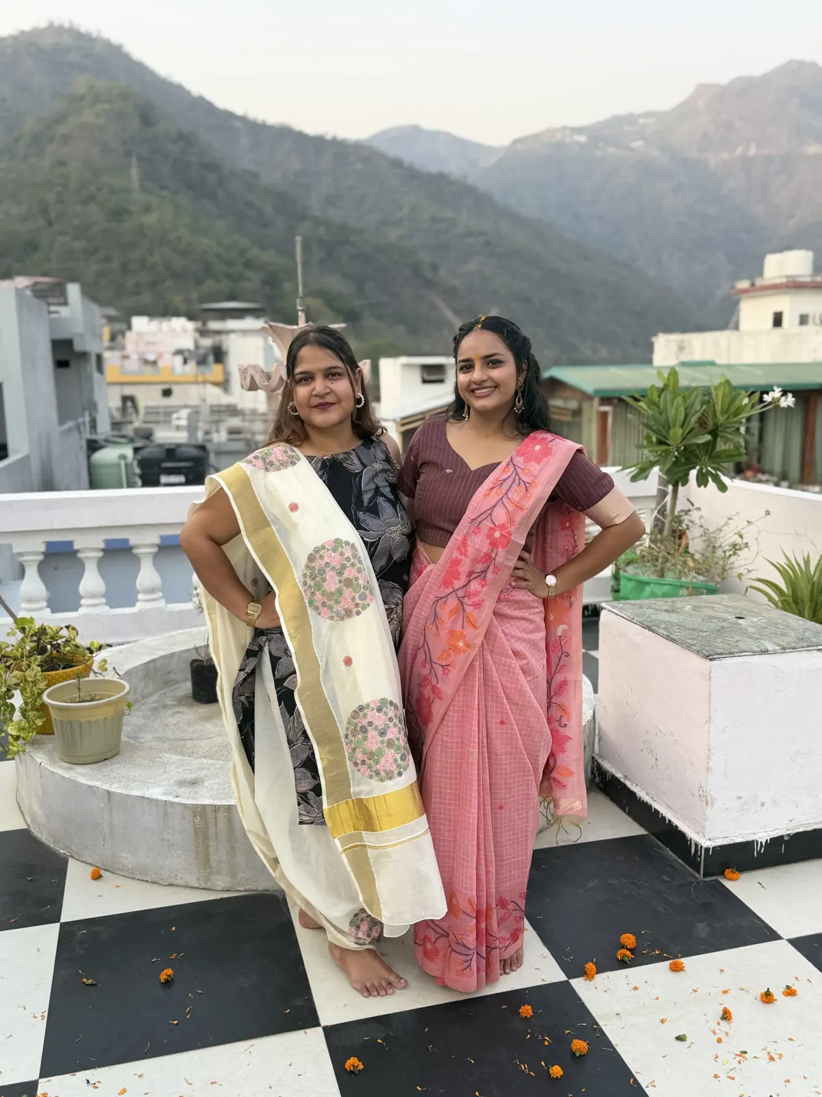 Two graduates in traditional Indian sarees on the rooftop terrace scattered with marigold petals after their yoga teacher training ceremony