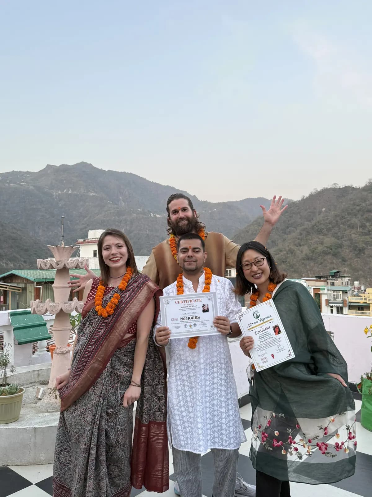 Four international graduates in marigold garlands celebrating with their certificates on the Rishikesh rooftop terrace