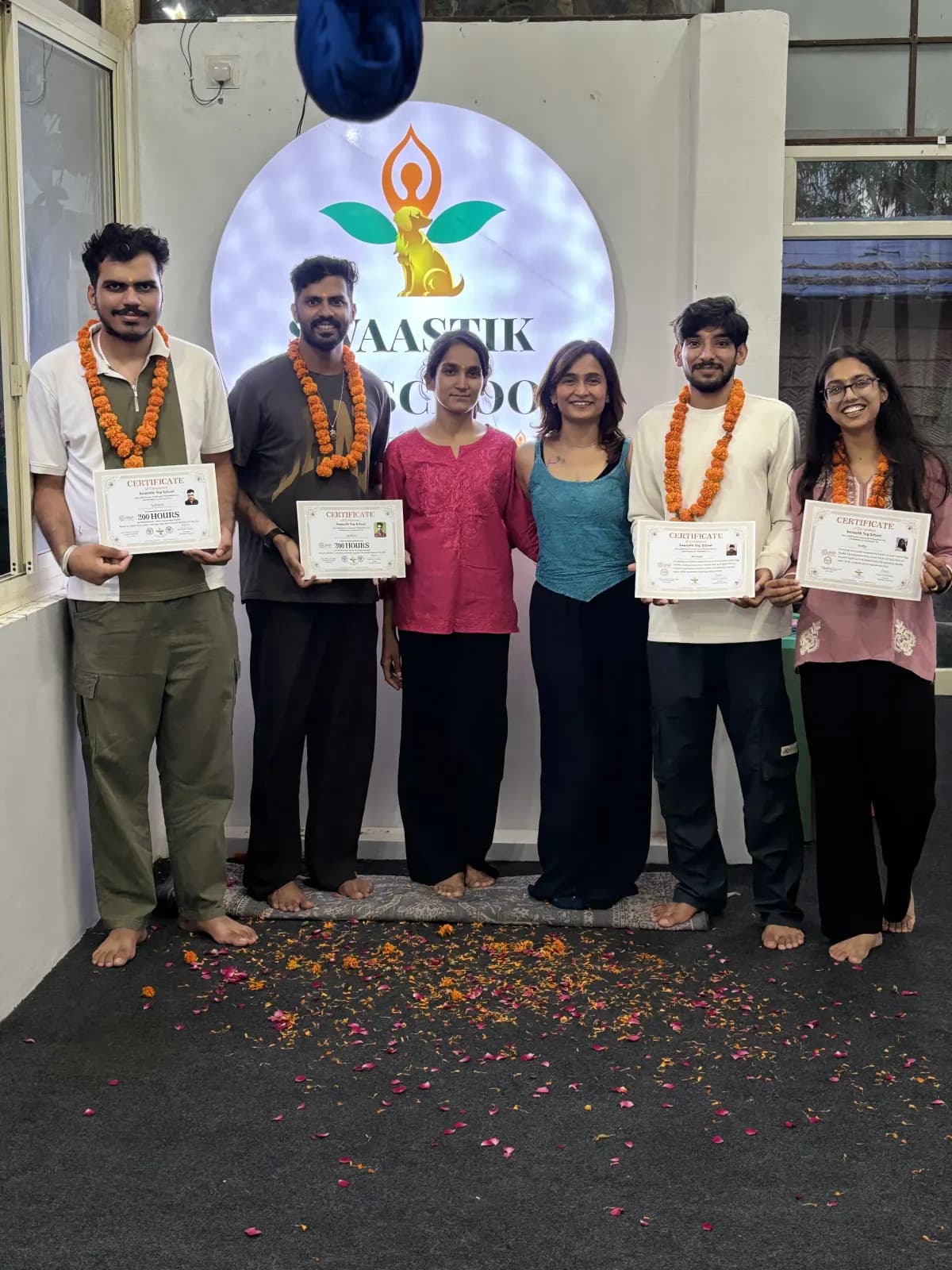 Men and women graduates in marigold garlands holding yoga teacher training certificates in front of the illuminated Swaastik Yog School logo