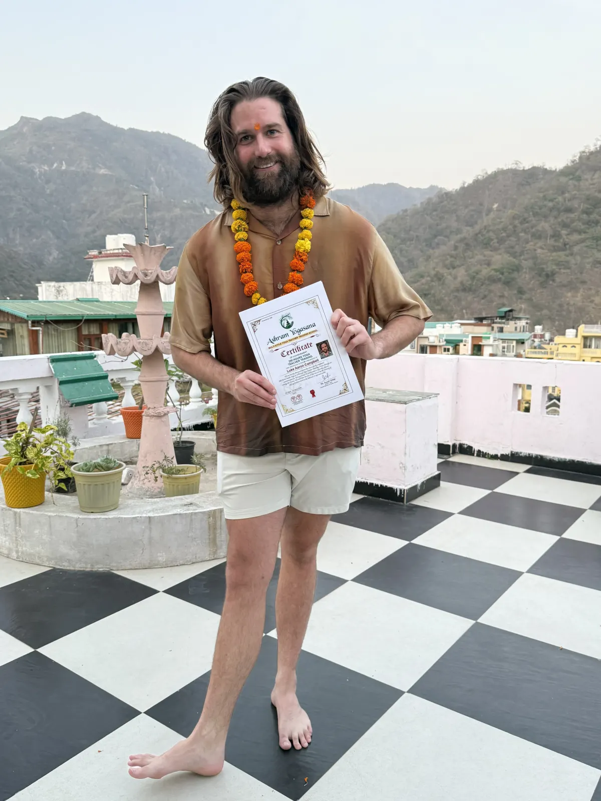 International male graduate standing barefoot with a marigold garland and his yoga teacher training certificate on the Rishikesh rooftop