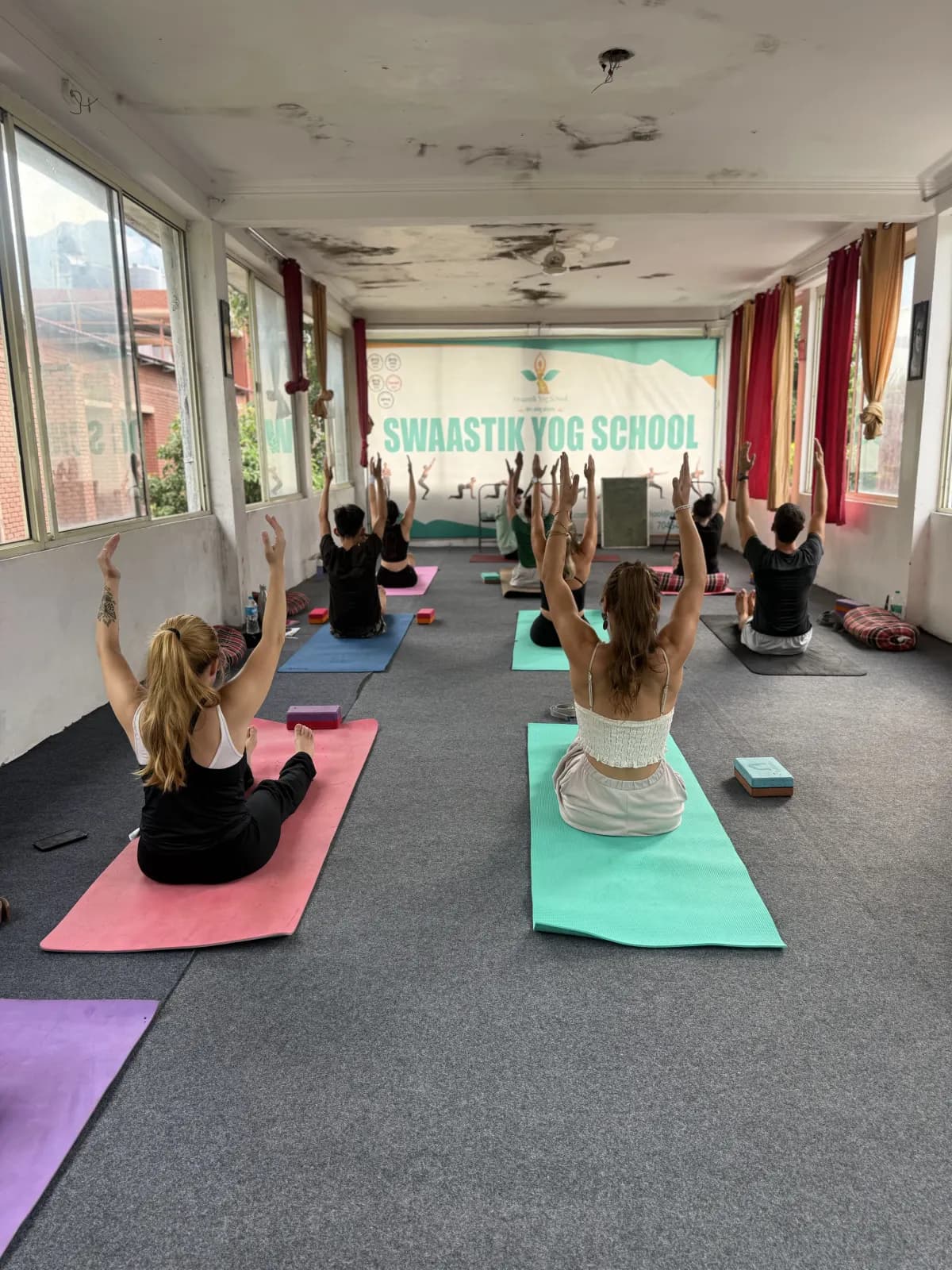 Students raising their arms in an asana class in the long yoga hall under the Swaastik Yog School banner