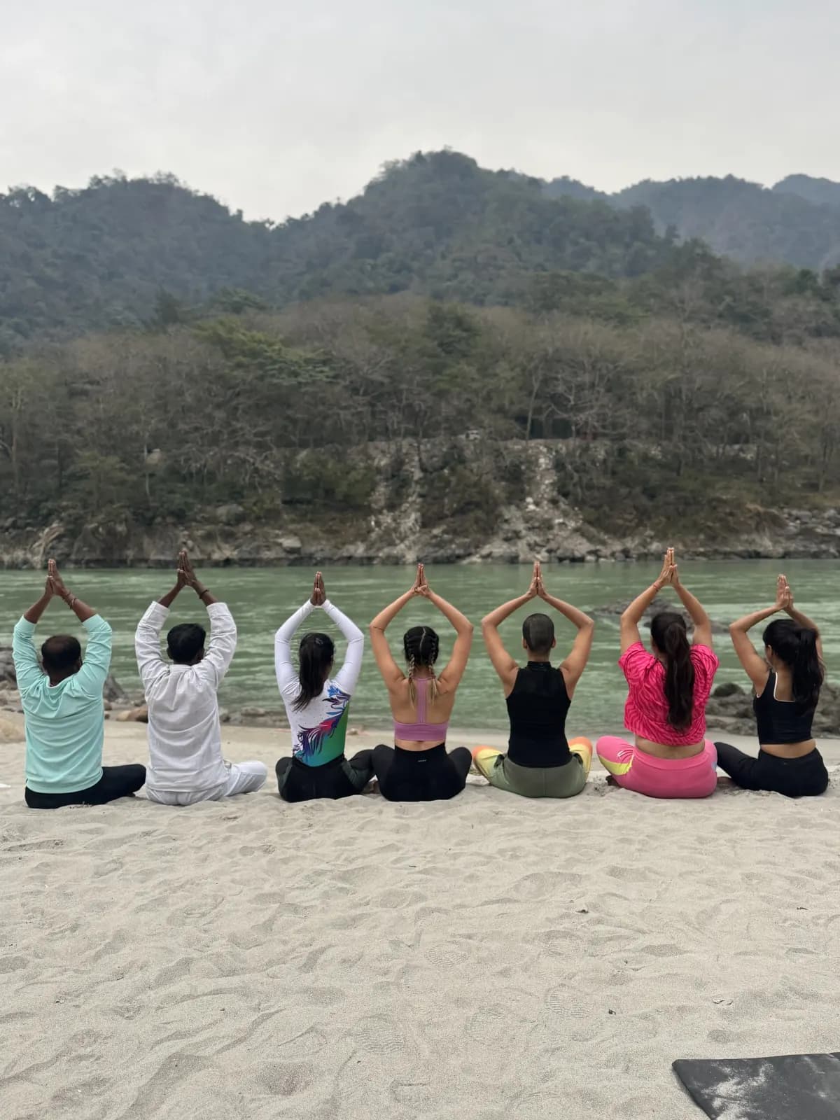 Students seated on the sand with arms raised in prayer, facing the Ganga and the forested hills of Rishikesh
