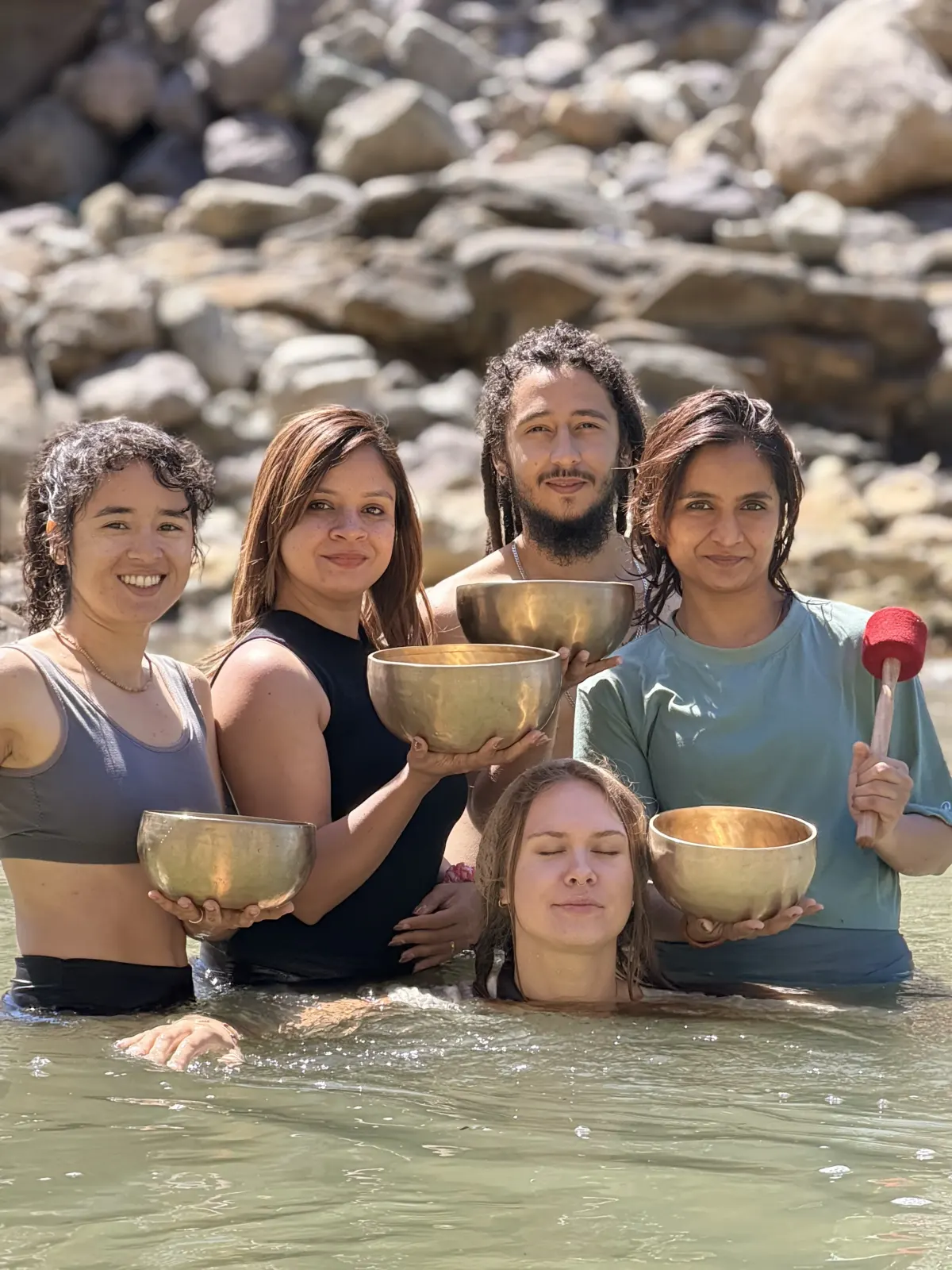 Sound healing students holding Tibetan singing bowls in the Ganga river as a classmate floats between them
