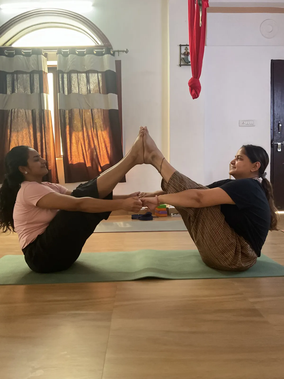 Two students in partner Boat pose (Navasana), hands and feet joined, during a yoga class