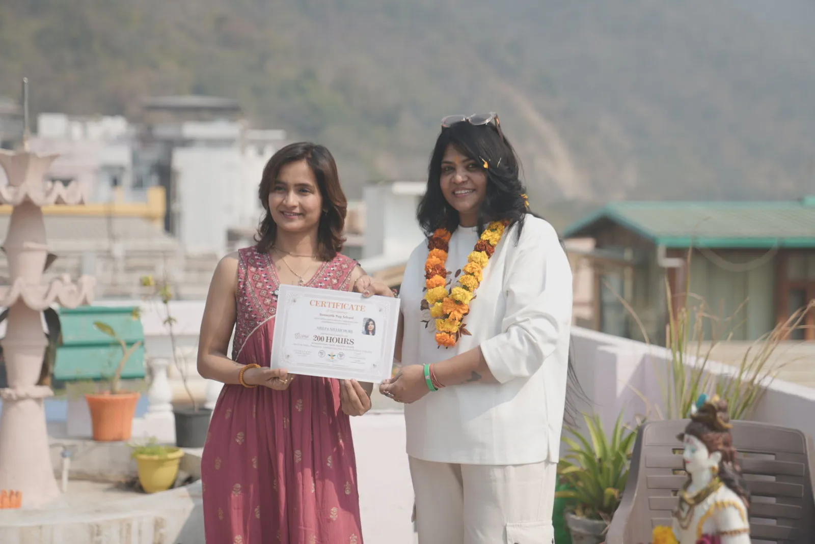 Founder Mayuri handing a 200 Hours Yoga Alliance certificate to a garlanded graduate on the Swaastik Yog School rooftop in Rishikesh