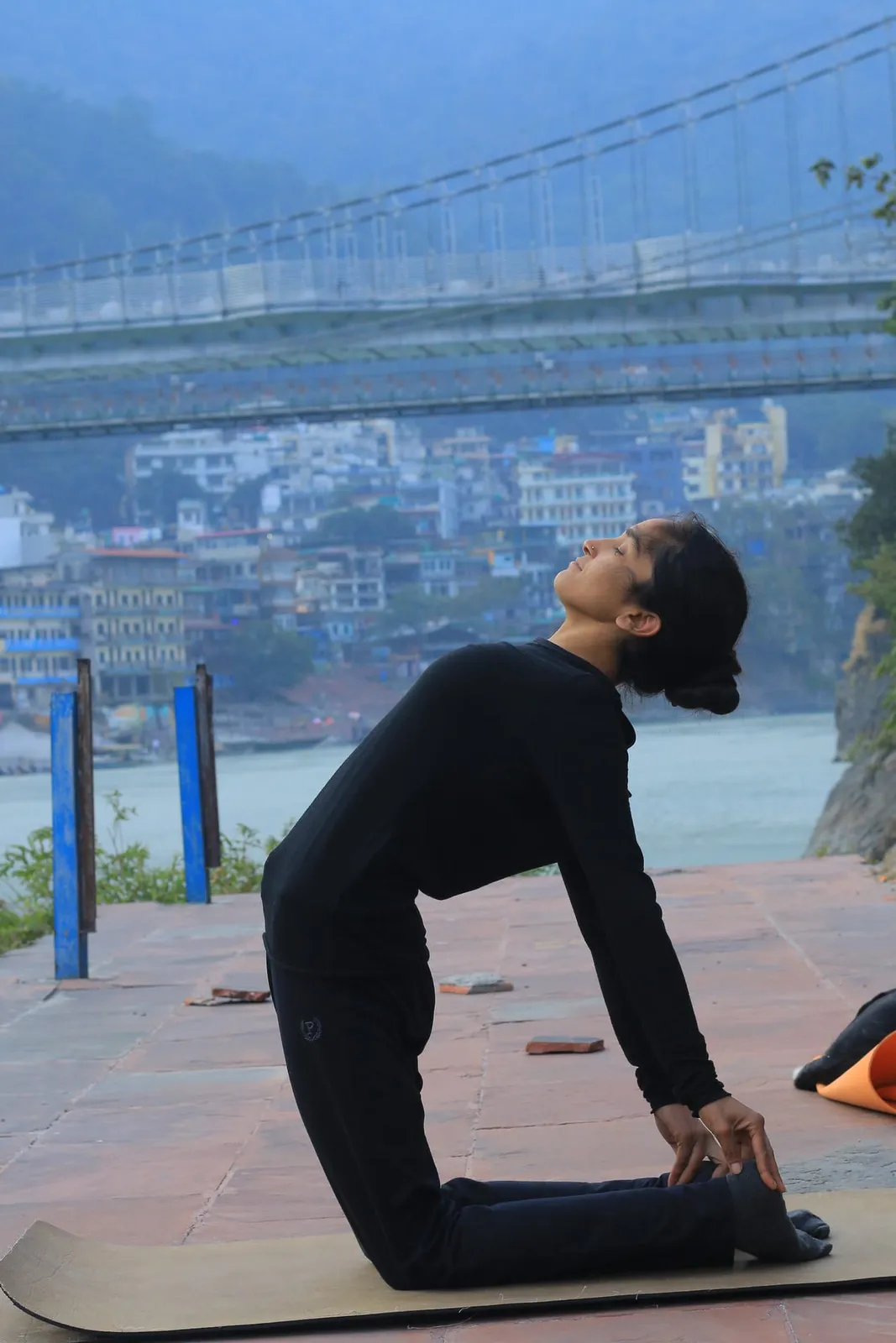 Yoga teacher practicing camel pose on the Ganges riverbank with Ram Jhula bridge and Rishikesh town behind