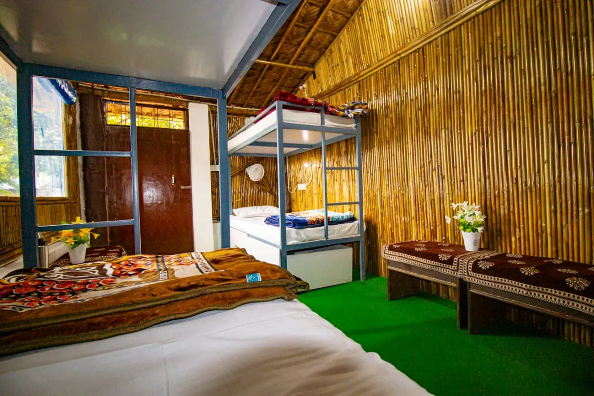 Inside the bamboo dorm cottage at Swaastik Yog School in Tapovan, Rishikesh, with a bunk bed, a single bed, a wall fan and a cushioned bench