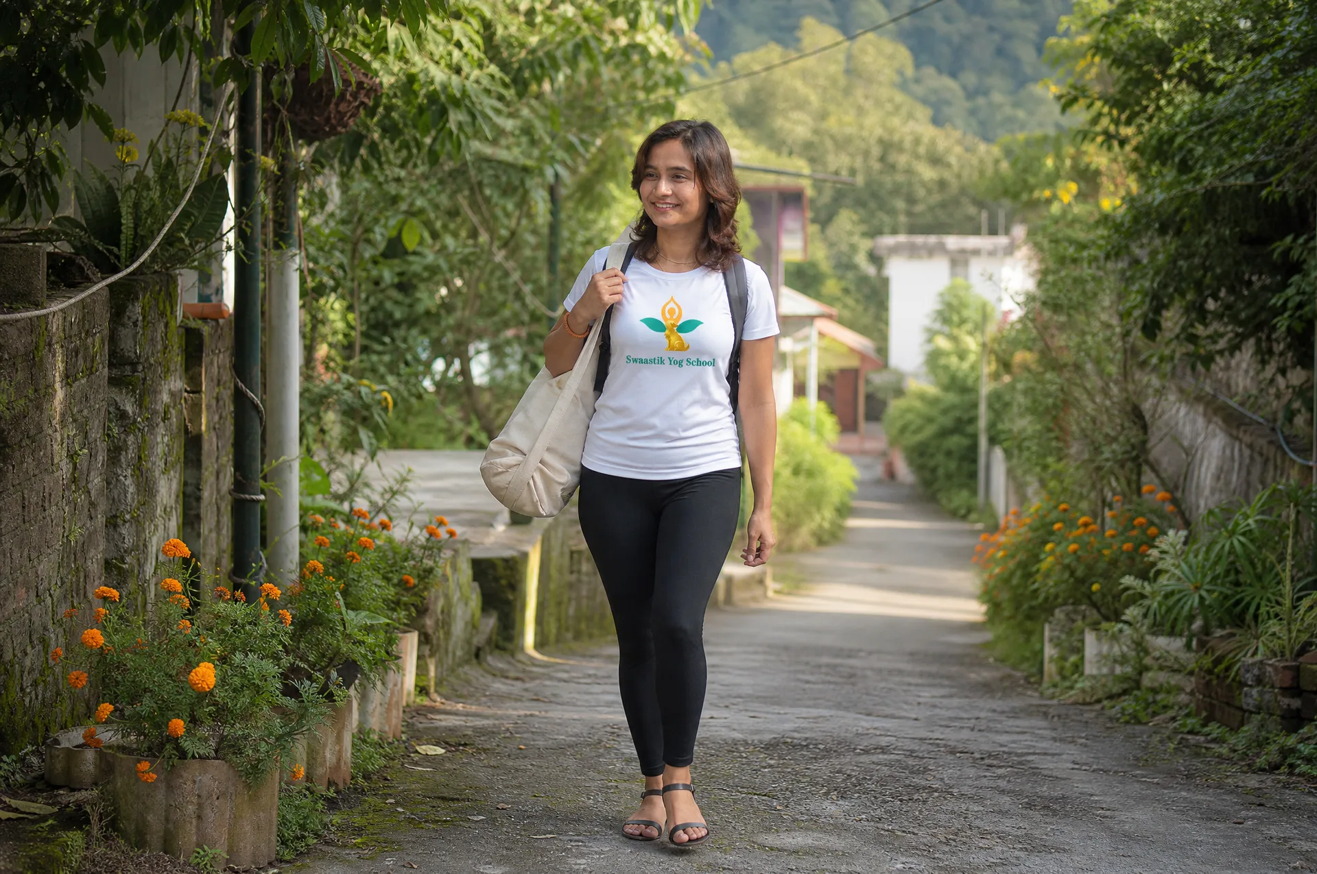 Mayuri walking down a leafy Tapovan lane in Rishikesh carrying a yoga mat bag over her shoulder