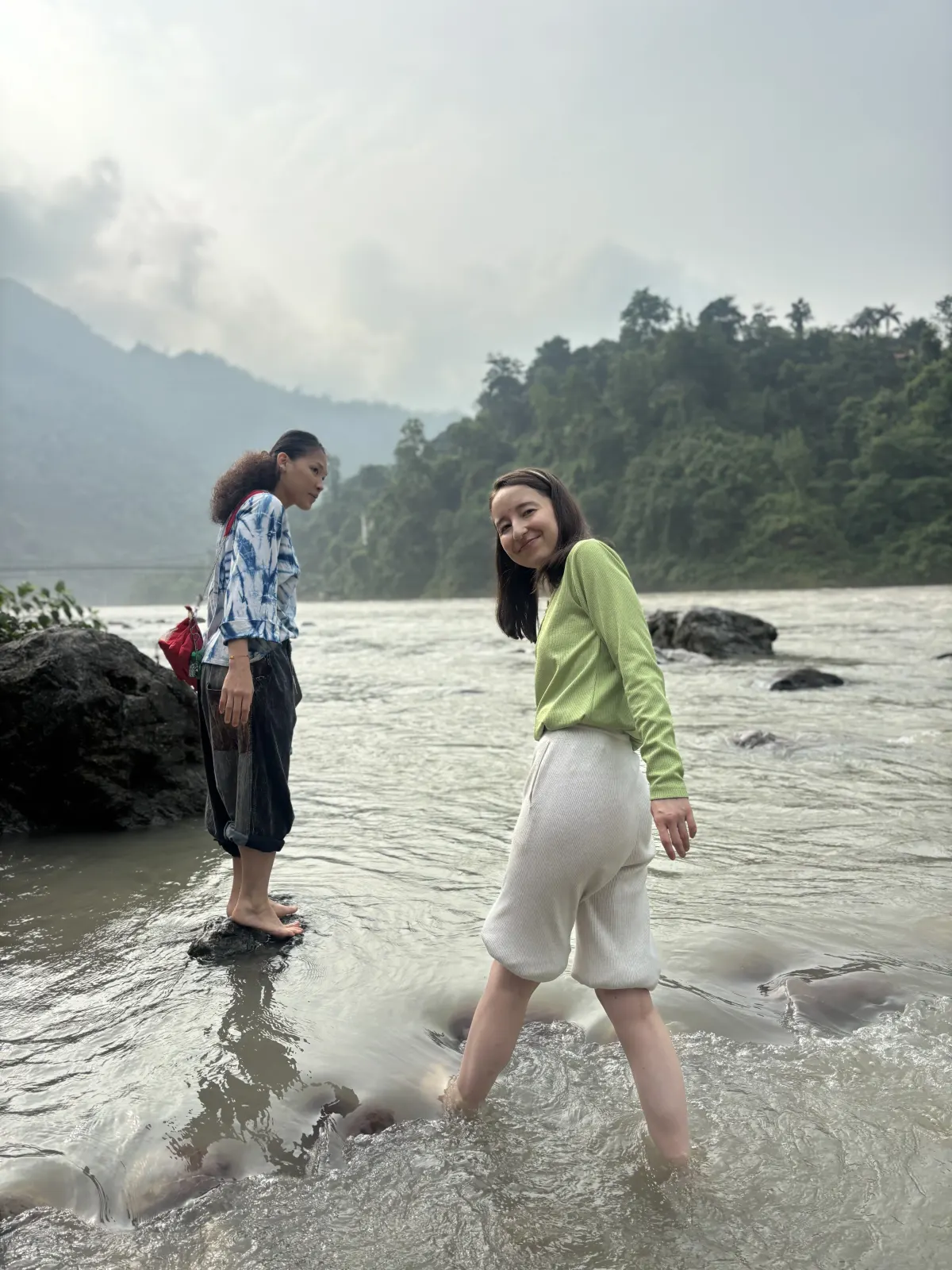 Two women wading at the edge of the Ganga on a cloudy day, with forested Himalayan foothills behind