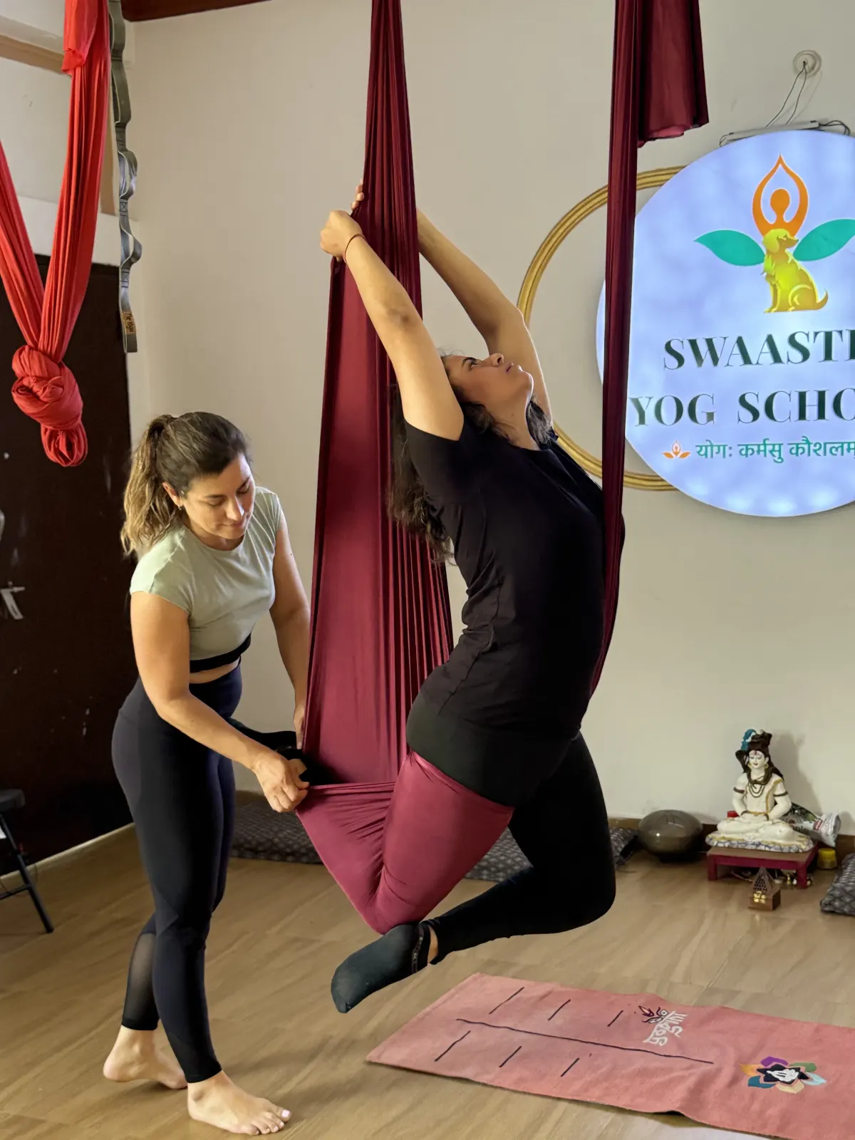 A woman steadies a student's leg in a burgundy aerial yoga hammock as the student arches back holding the fabric, in a studio with the Swaastik Yog School logo on the wall