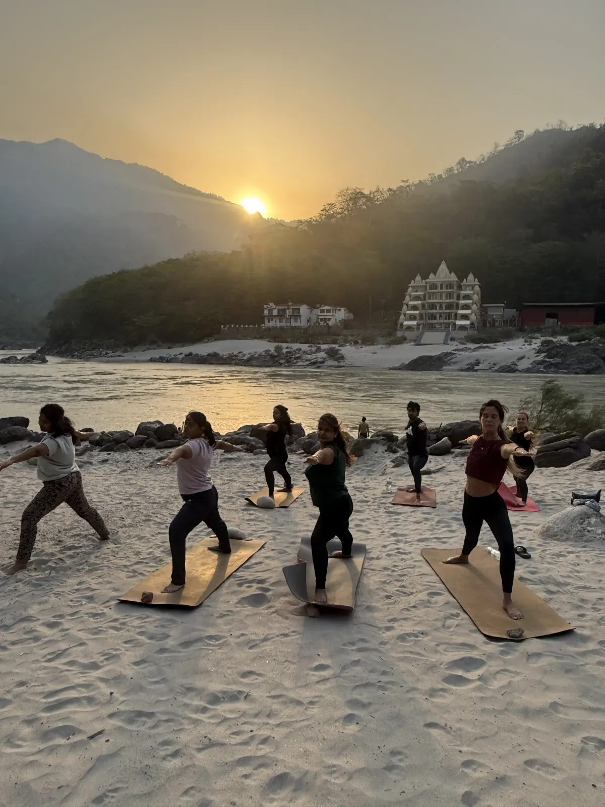 Students in warrior pose on a white-sand Ganga beach near Tapovan at sunset, with a temple and forested hills across the river