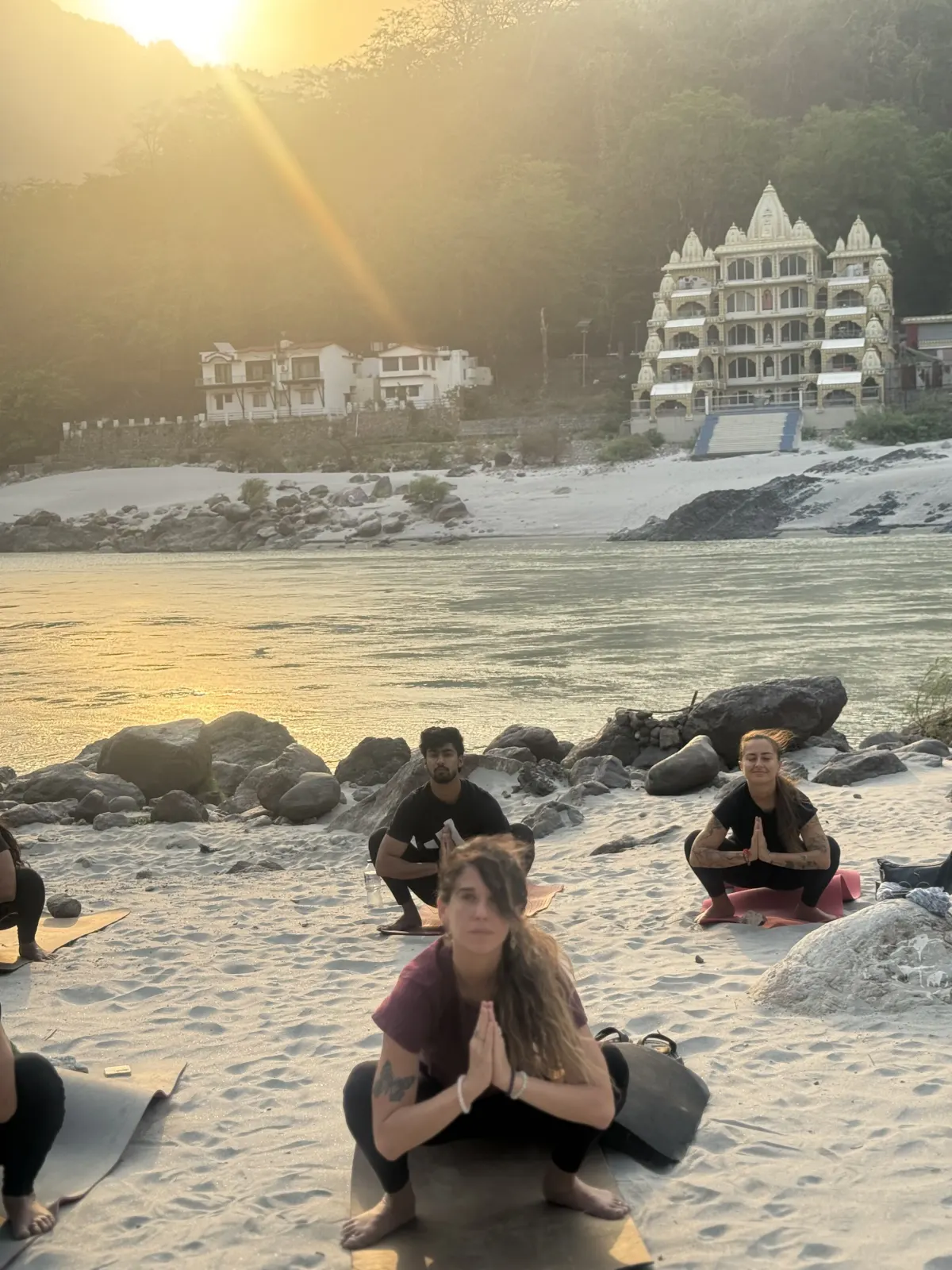 Yoga teacher training students in a deep squat with hands in prayer on the Ganga beach in Rishikesh, in golden light