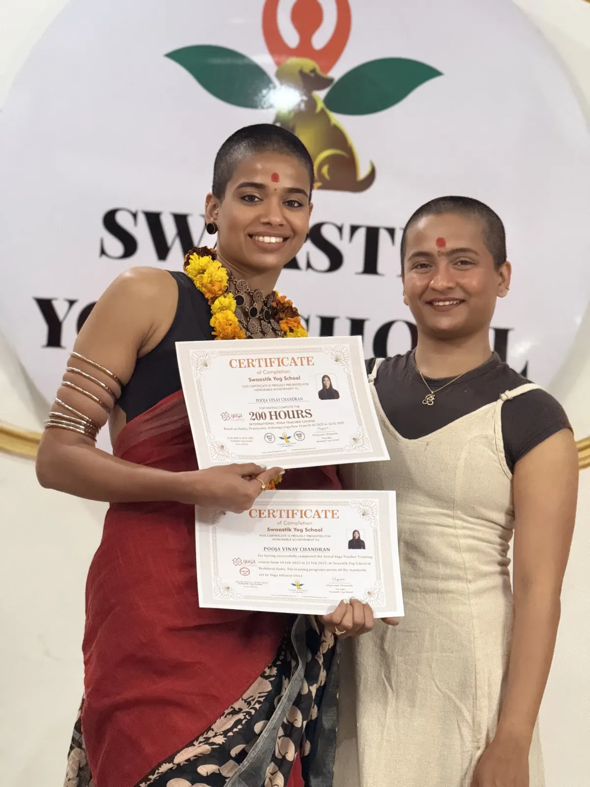 Graduate in a red saree and marigold garland holding her Swaastik Yog School 200 hours and aerial yoga certificates, standing beside a fellow student