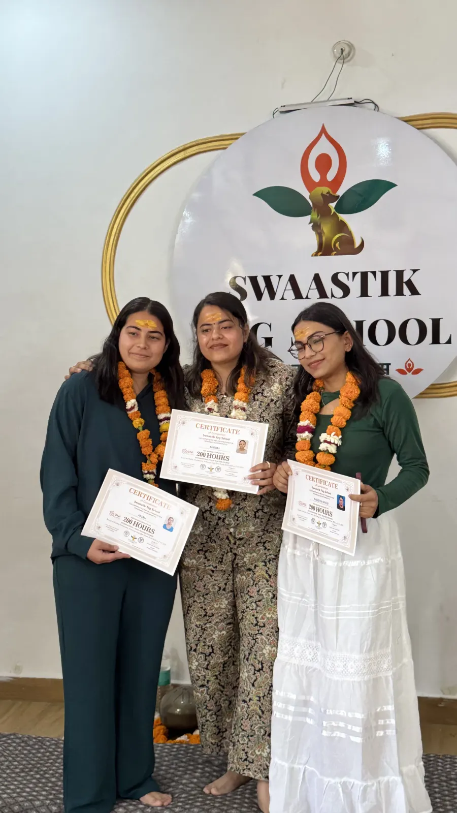Three women wearing orange marigold garlands and tilak marks hold their 200-hour certificates in front of the Swaastik Yog School logo at their graduation ceremony
