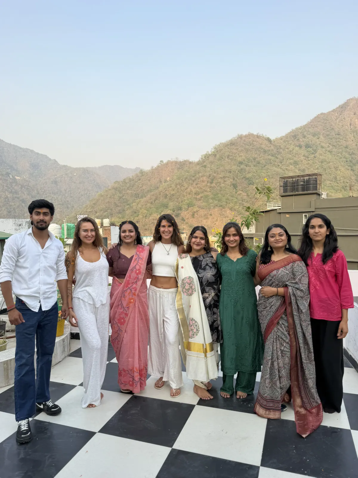 A graduating yoga teacher training batch in traditional dress on the Swaastik Yog School rooftop in Tapovan, Rishikesh, with green hills behind