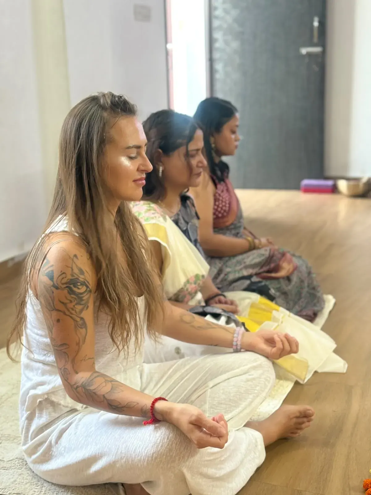 Tre studentesse della Swaastik Yog School sedute a gambe incrociate in fila, con gli occhi chiusi e le mani in una mudra, durante una lezione di meditazione a Rishikesh